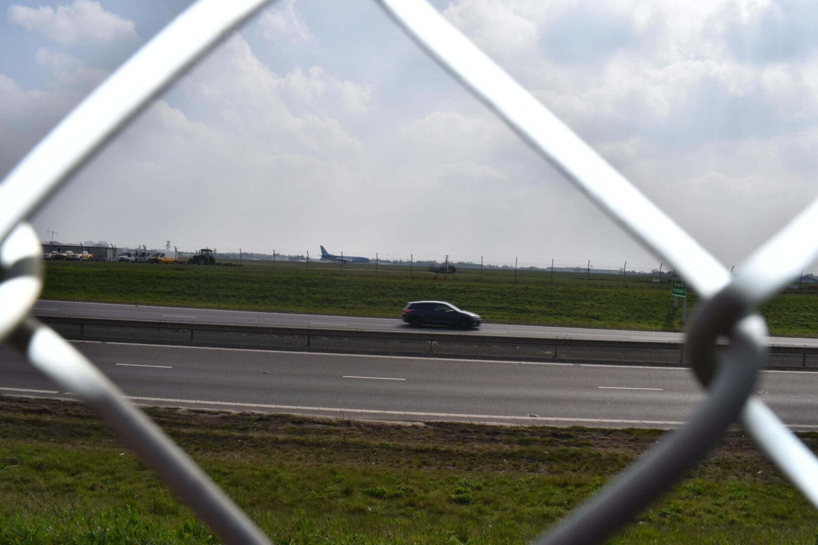 Car on highway seen through chain-link fence.